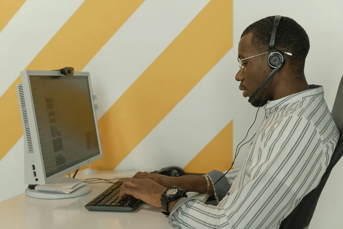 A person wearing a headset works on a computer at a desk, set against a yellow and white striped wall. The atmosphere is focused and professional.