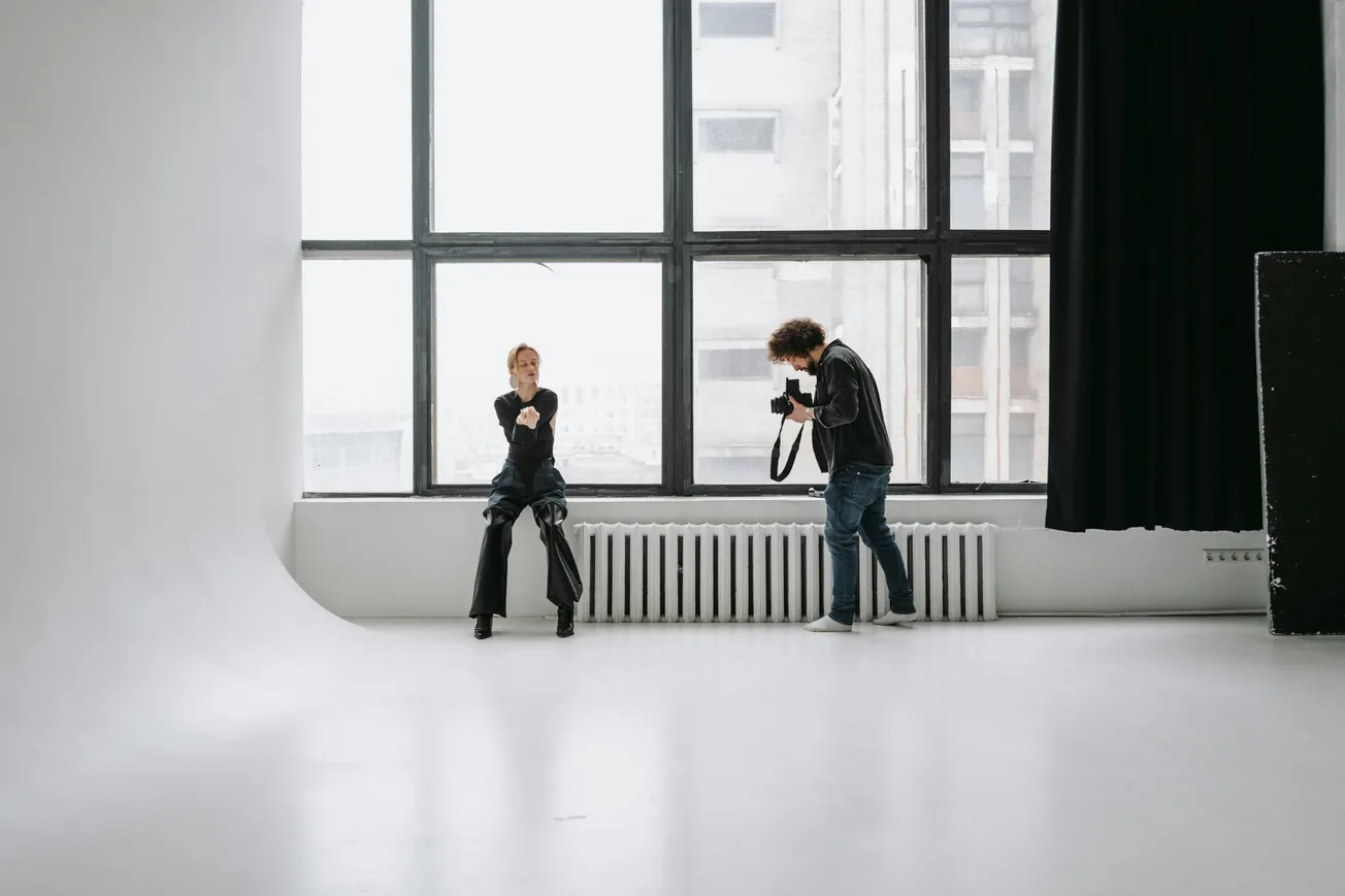 Photographer taking a picture of a seated person in a bright studio. The room features large windows and a minimalist, modern aesthetic.