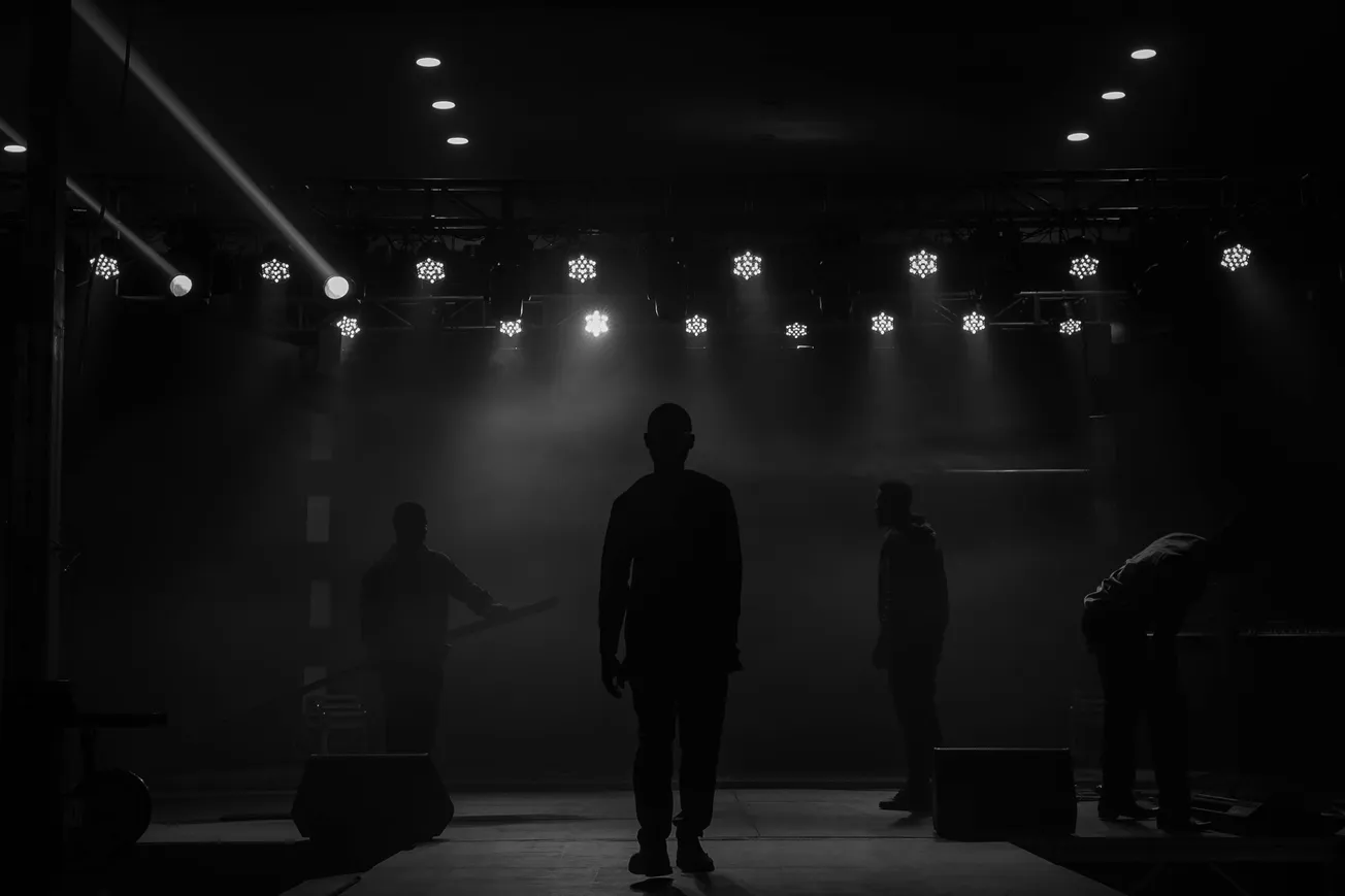 A silhouetted man stands center stage under bright spotlights, surrounded by stage crew in a dimly lit concert hall, evoking anticipation and focus.