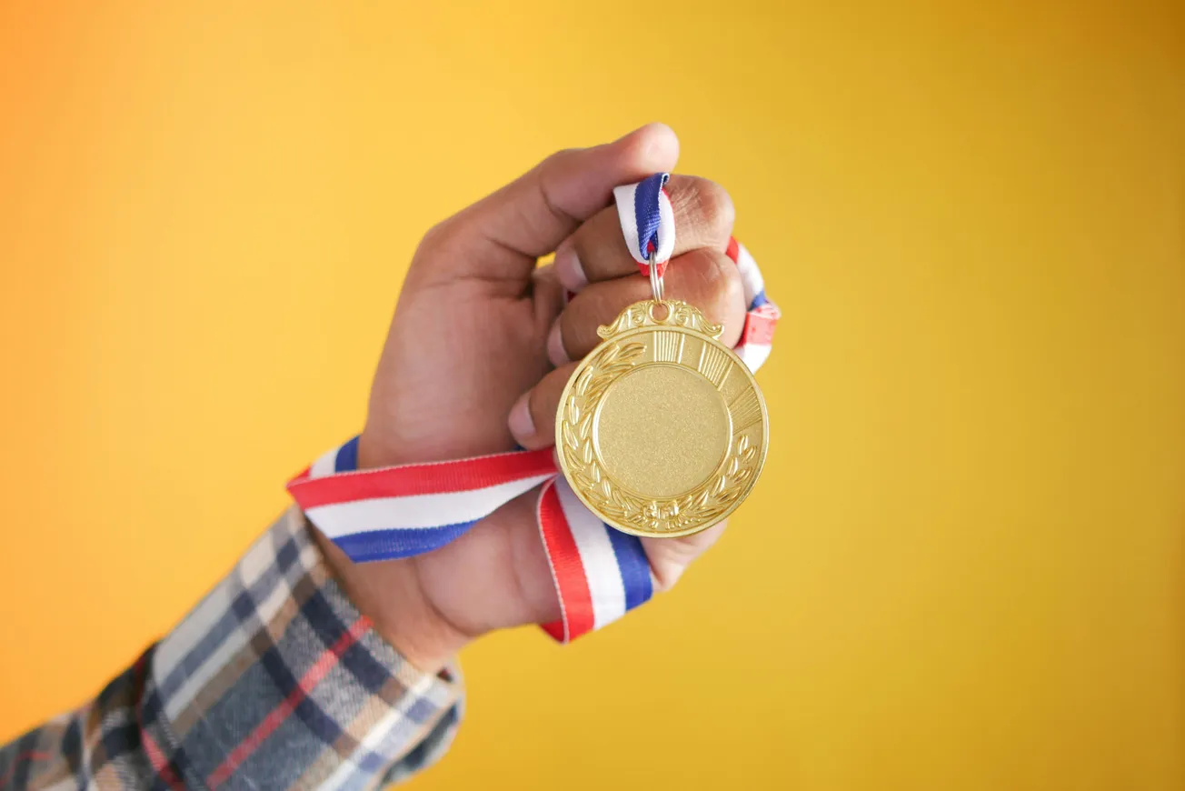 A hand holding a gold medal with a red, white, and blue ribbon against a yellow background, conveying achievement and pride.