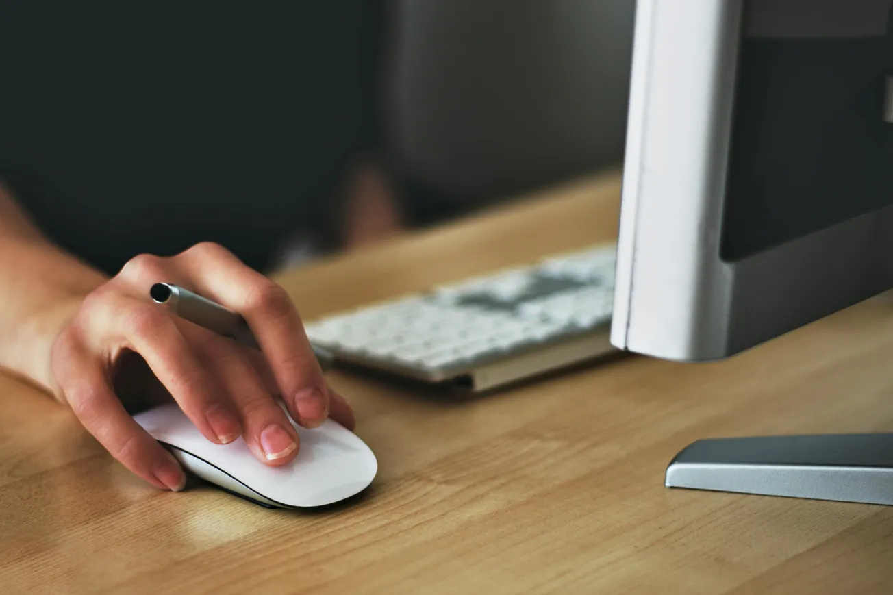 A hand holding a pen rests on a computer mouse beside a keyboard and monitor on a wooden desk, suggesting focus and productivity in a workspace.