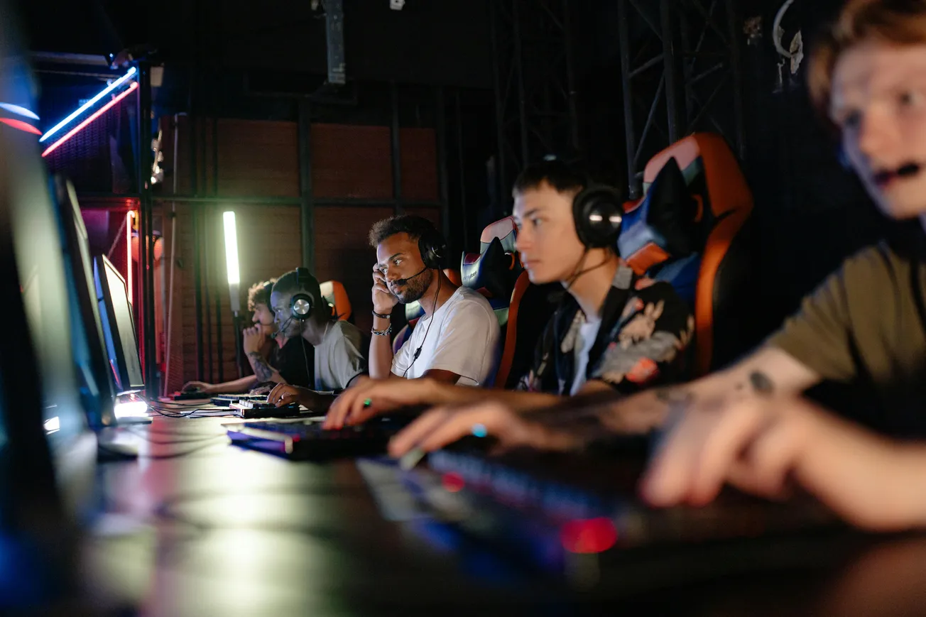 Four focused gamers wearing headsets sit in a dimly lit room, intensely playing on computers. Neon lights create an energetic, competitive atmosphere.