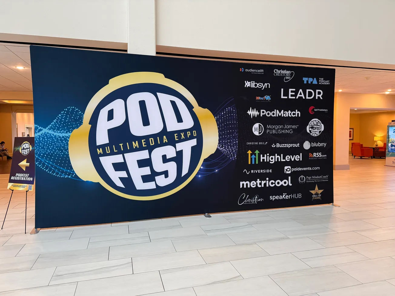Large banner for PODFEST Multimedia Expo featuring the event name in bold letters with headphone imagery. Surrounding are logos of various sponsors, set in a spacious hallway.