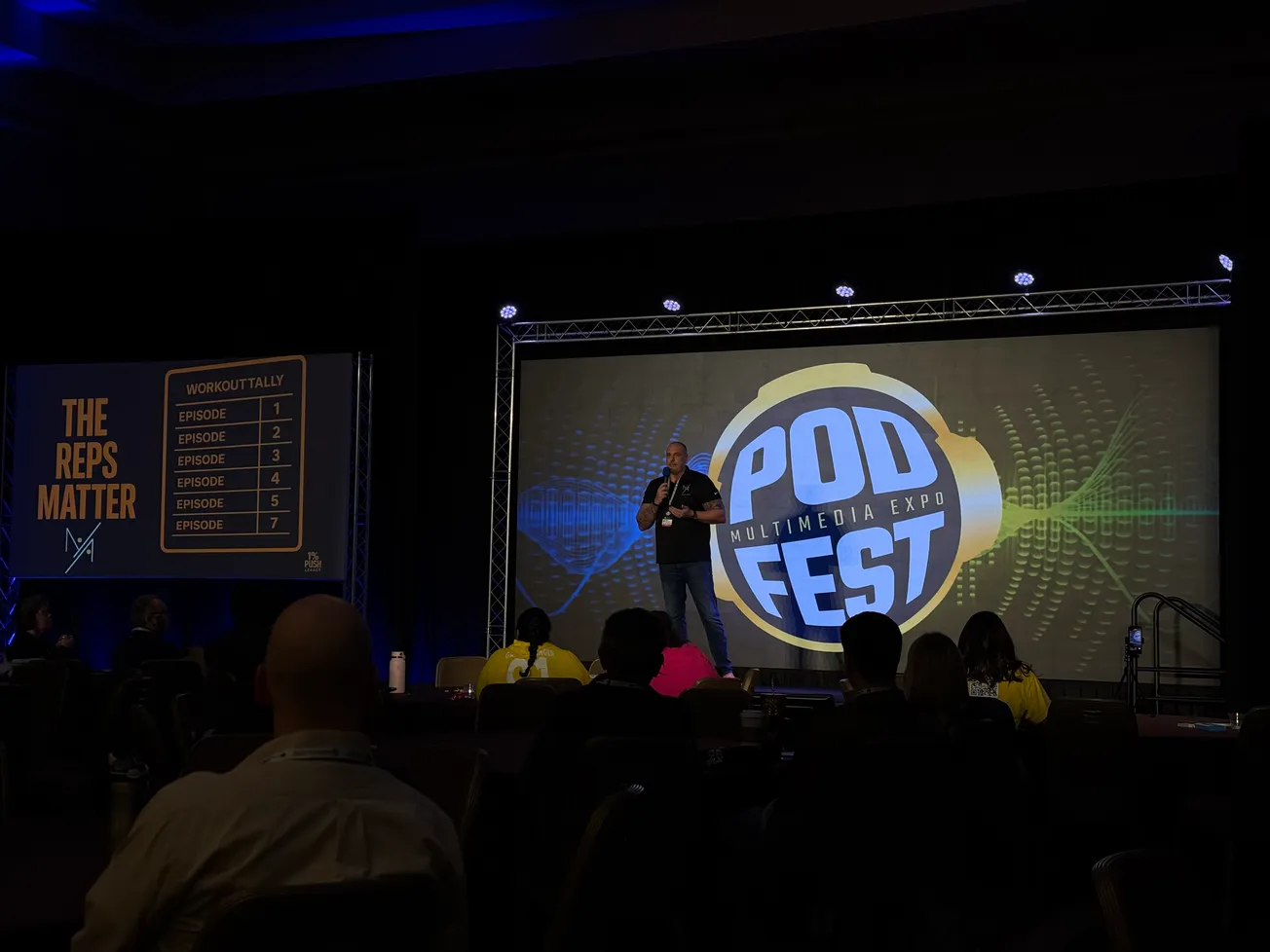 A speaker on stage at Pod Fest Multimedia Expo, with a large screen displaying event graphics and the text, "The Reps Matter" and episode list. Audience seated in dimly lit room.