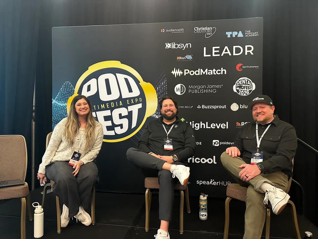 Three people seated in front of a "Podfest Multimedia Expo" backdrop. They are smiling and wearing conference badges. The mood is friendly and relaxed.