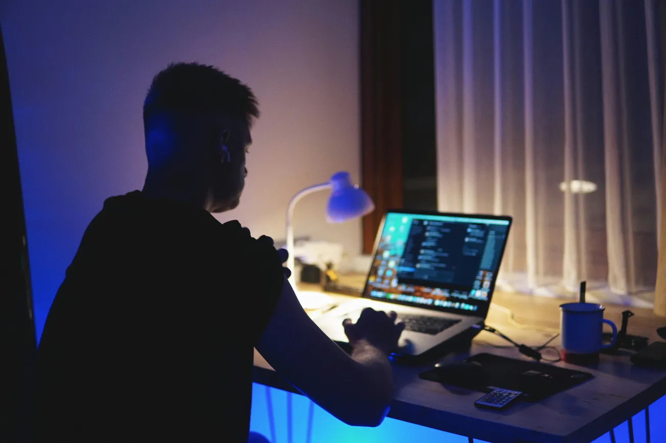 A person sits in a dimly lit room working on a laptop, with code visible on the screen. A desk lamp casts a soft glow, creating a focused ambiance.