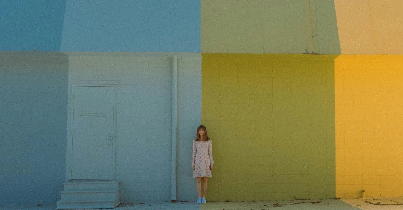 A woman in a pale dress stands against a large wall divided into blue and yellow sections. The minimalist setting conveys a calm, abstract feel.