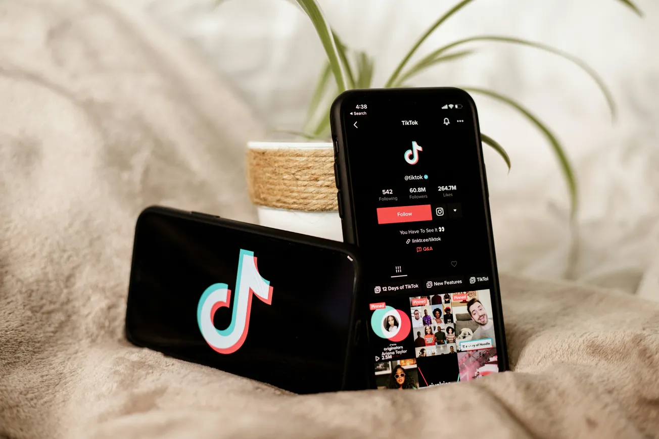 Two smartphones on a soft beige blanket. One shows the TikTok app's feed; the other displays the TikTok logo. A small plant in a pot is in the background.