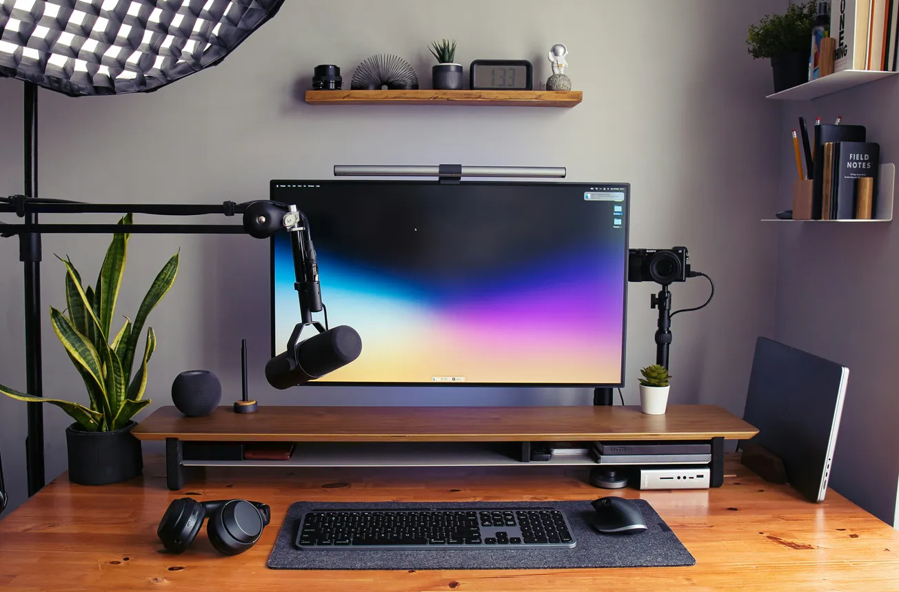 Modern workspace with a large monitor displaying a gradient screensaver, microphone, headphones, green plants, and organized shelves, conveying a clean, creative vibe.