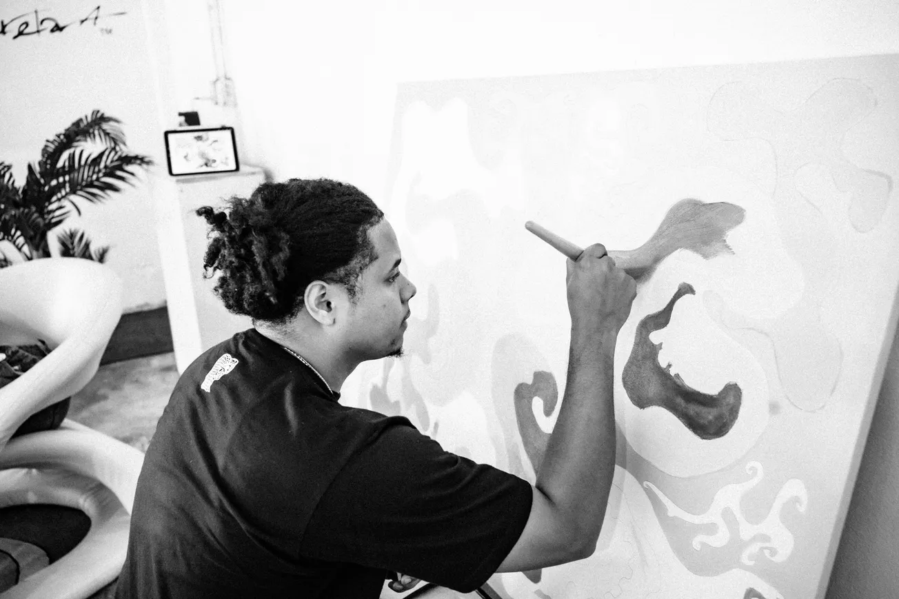 A person with curly hair is painting abstract shapes on a canvas in a bright studio. The scene conveys focus and creativity. Black and white photo.