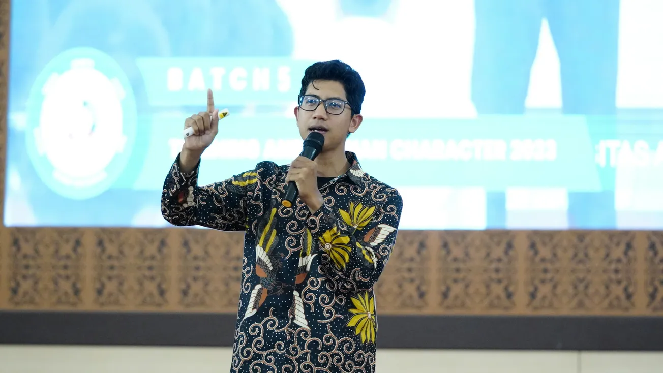 A man in a floral batik shirt holds a microphone, speaking energetically. Behind him, a blurred presentation screen with text enhances the formal setting.