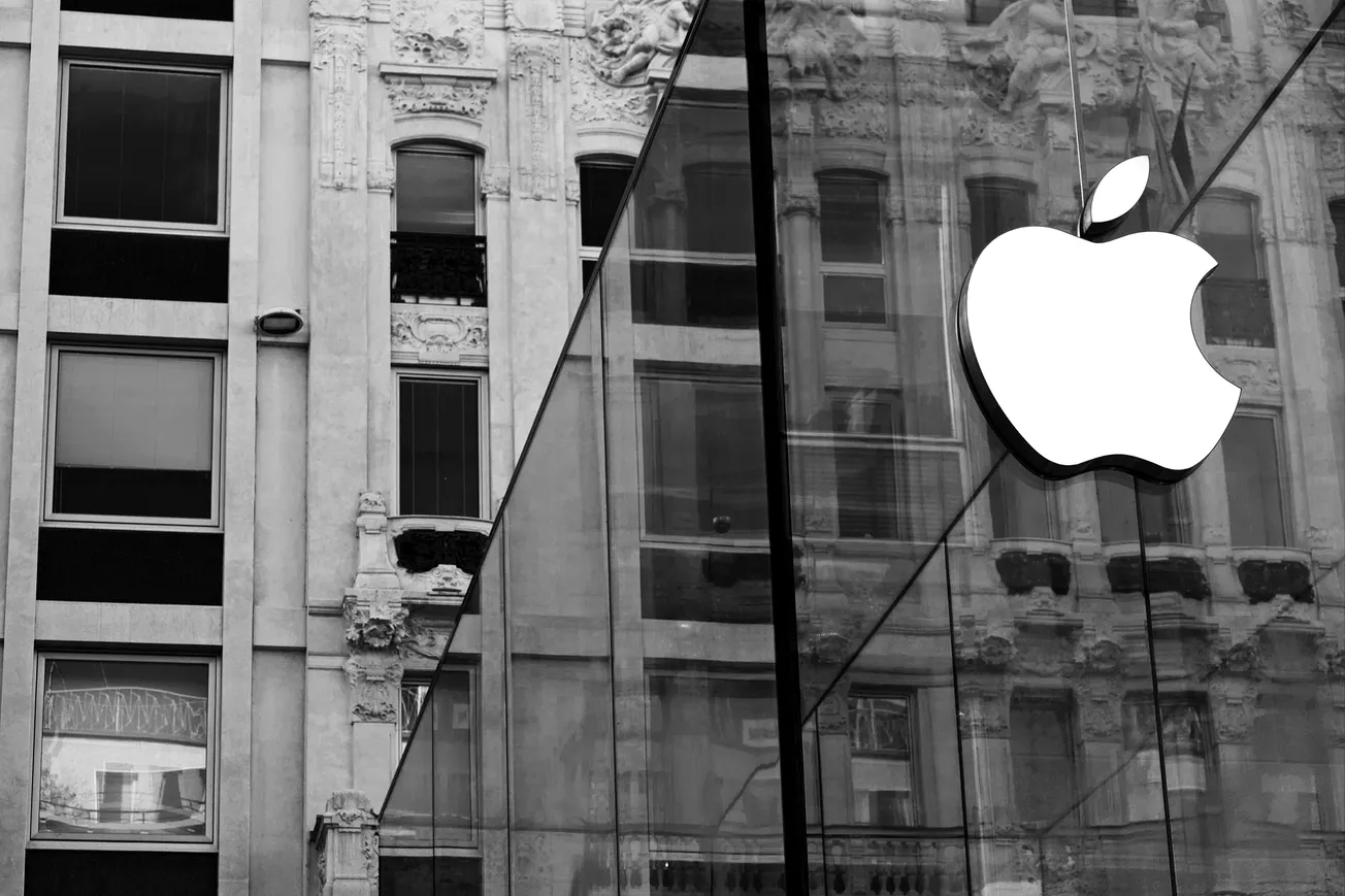Black and white image of a modern glass building with a white Apple logo, reflecting an ornate historic facade. The tone is elegant and timeless.