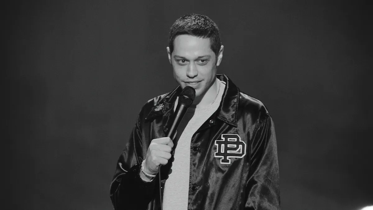 Pete Davidson wearing a black satin jacket holds a microphone, smiling slightly, with a neutral background. The scene conveys a casual, comedic tone.