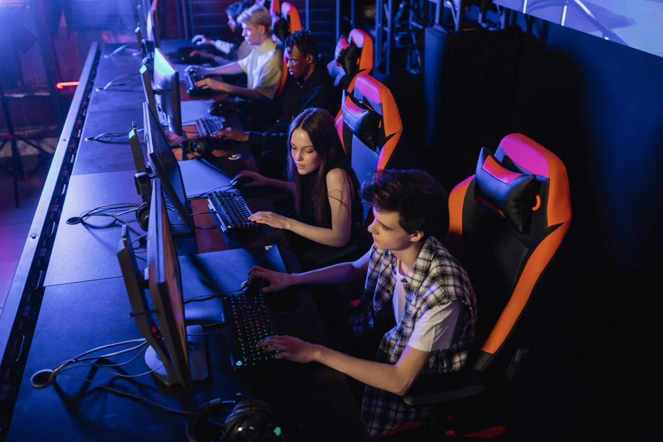 A group of focused gamers sit in a dimly lit room, each at a computer station with colorful RGB keyboards. They are seated in black and orange gaming chairs.