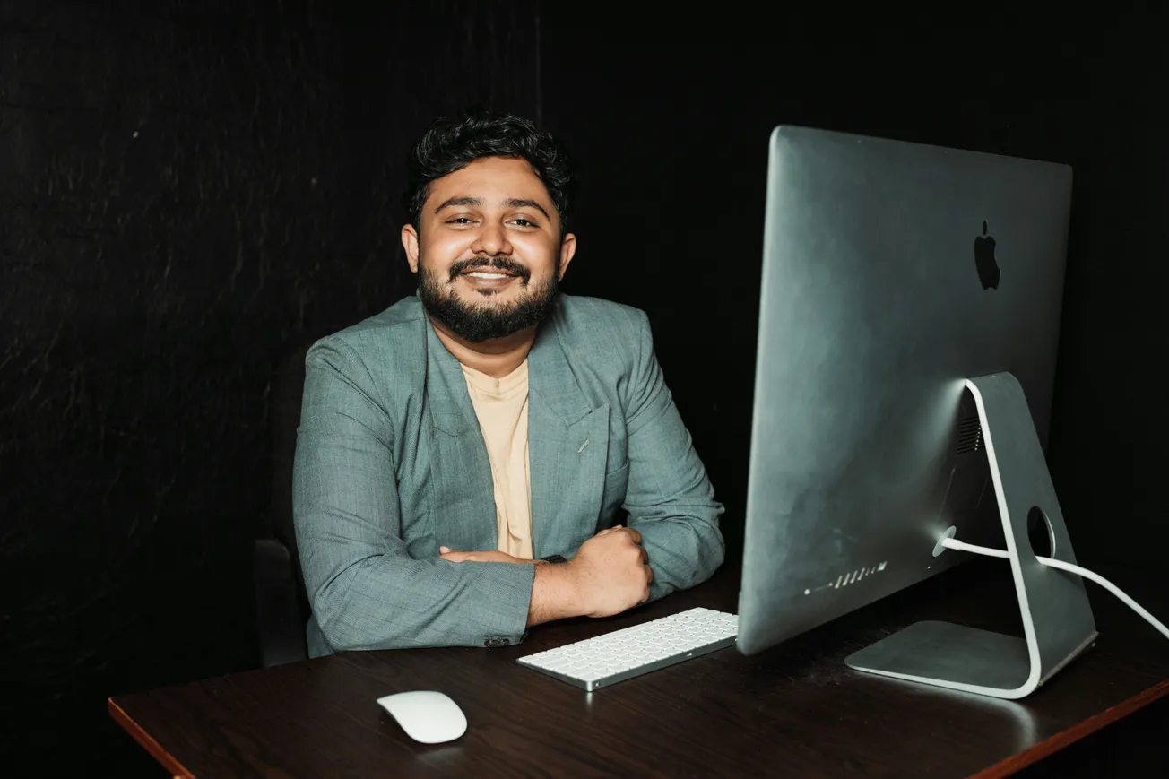 A person with a beard smiles warmly at a desk with a large computer, keyboard, and mouse against a dark background, creating a professional yet relaxed atmosphere.