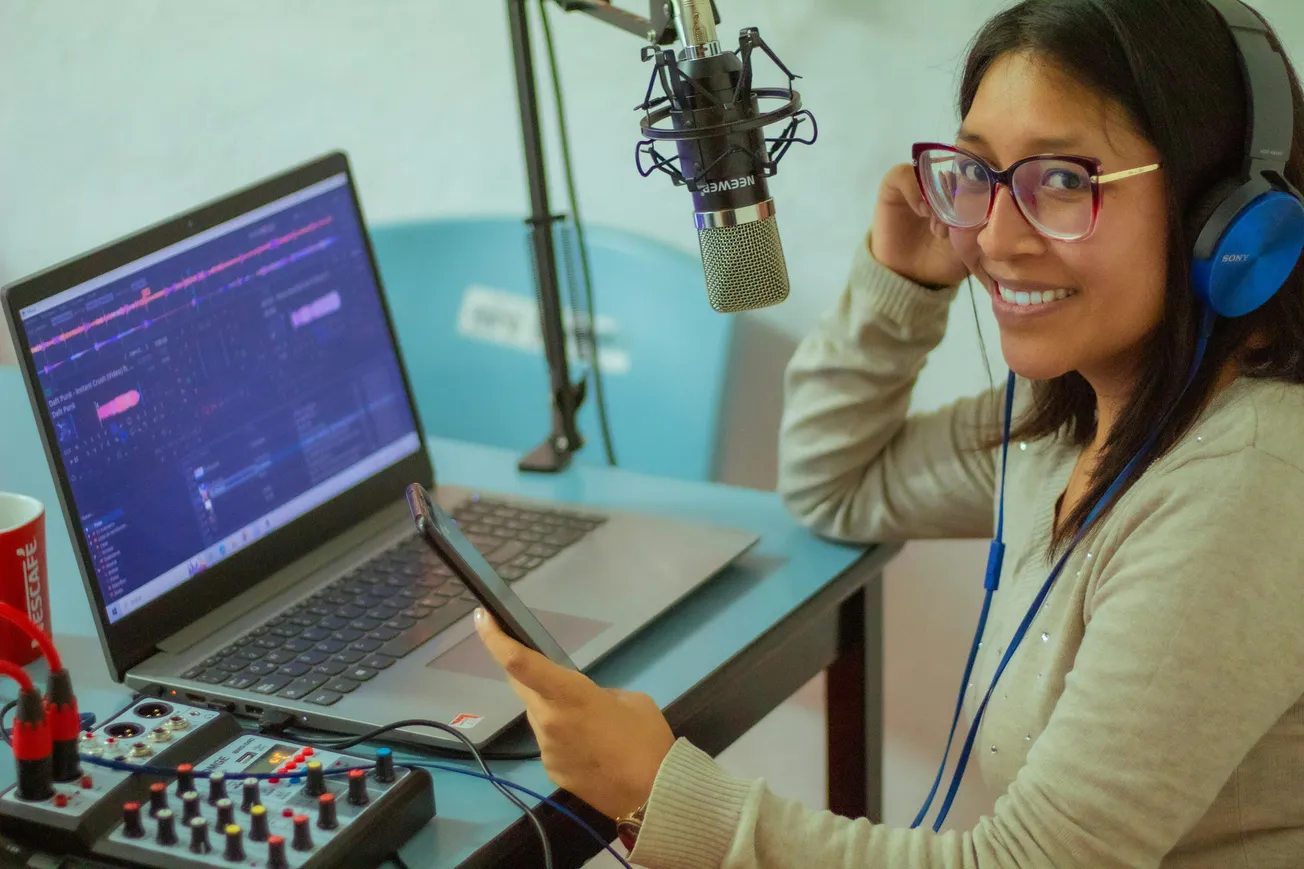 A woman smiling at a desk with a laptop, microphone, and mixer. She wears headphones and holds a smartphone, creating a cheerful podcasting vibe.