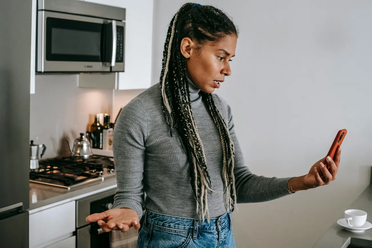 A woman with braided hair in a gray sweater looks at her phone with a curious expression in a modern kitchen. A teapot and cup are on the counter.