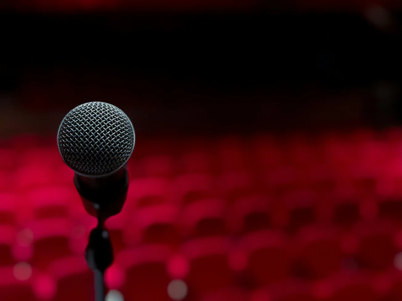 Close-up of a microphone on a stand, centered against a blurred background of empty red theater seats, evoking anticipation and solitude.