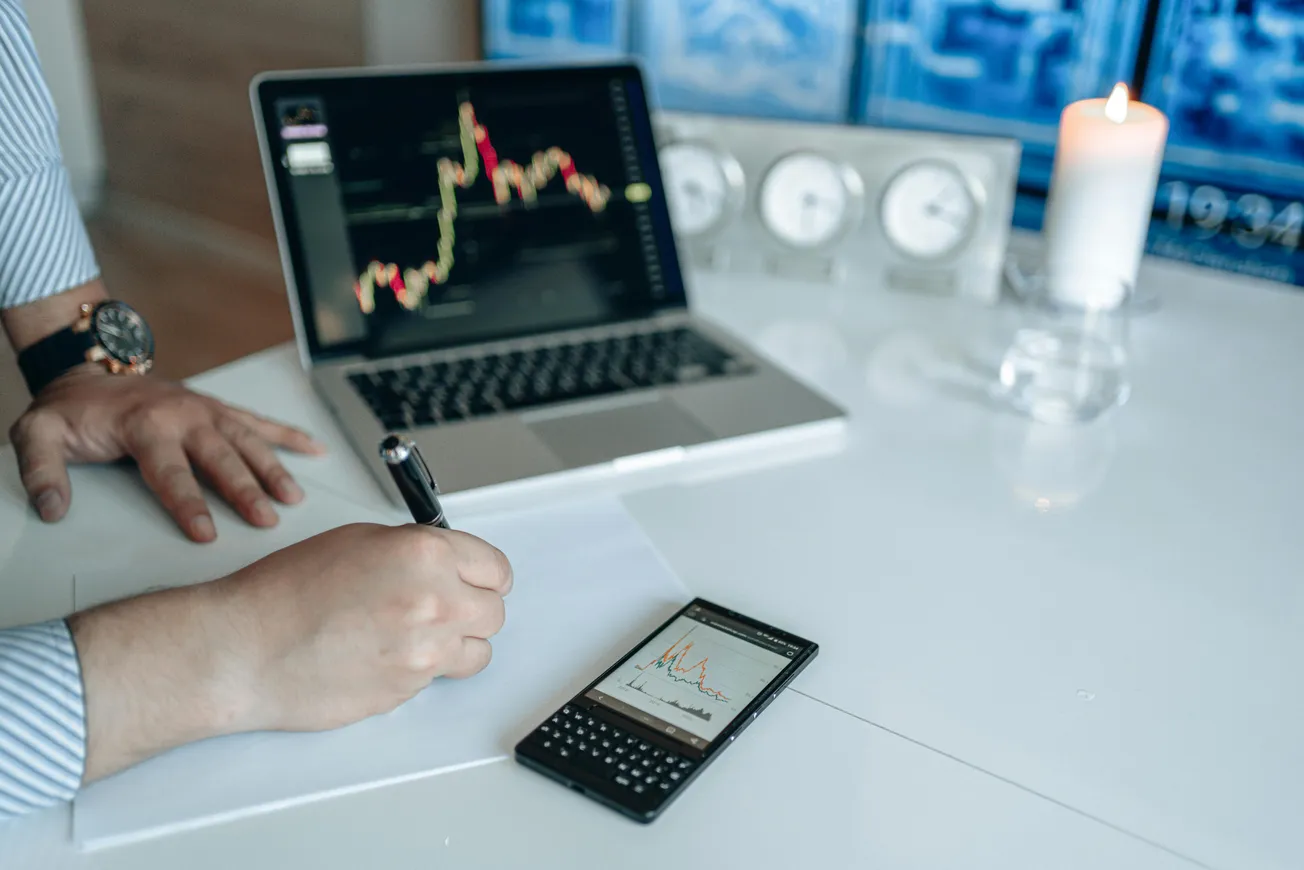 A person writes on paper at a desk, with a laptop showing stock charts and a smartphone displaying graphs. A lit candle adds a calm atmosphere.