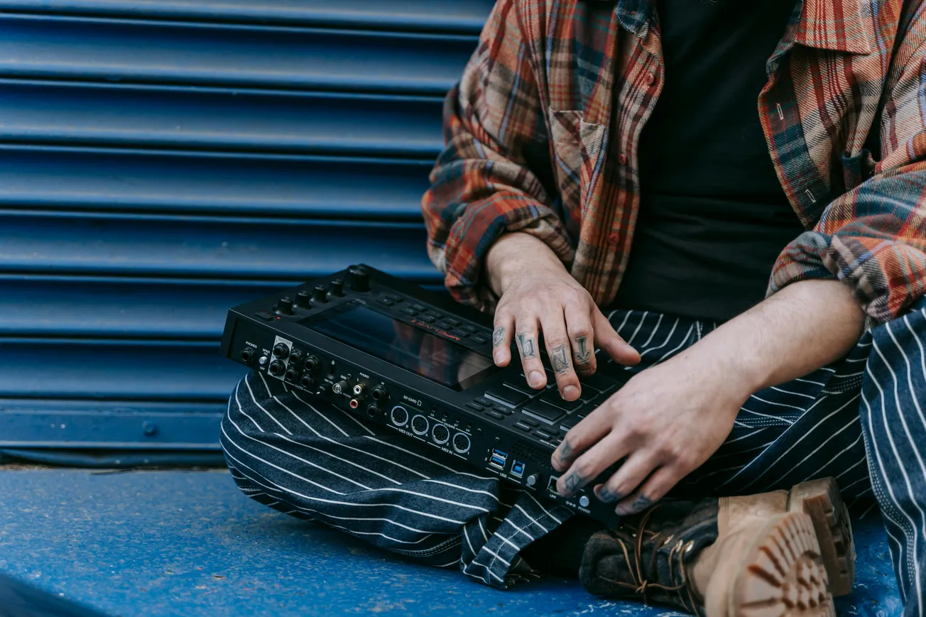 Person seated on the ground, holding an electronic music device. They're in casual plaid and striped clothing, next to a blue corrugated metal wall.