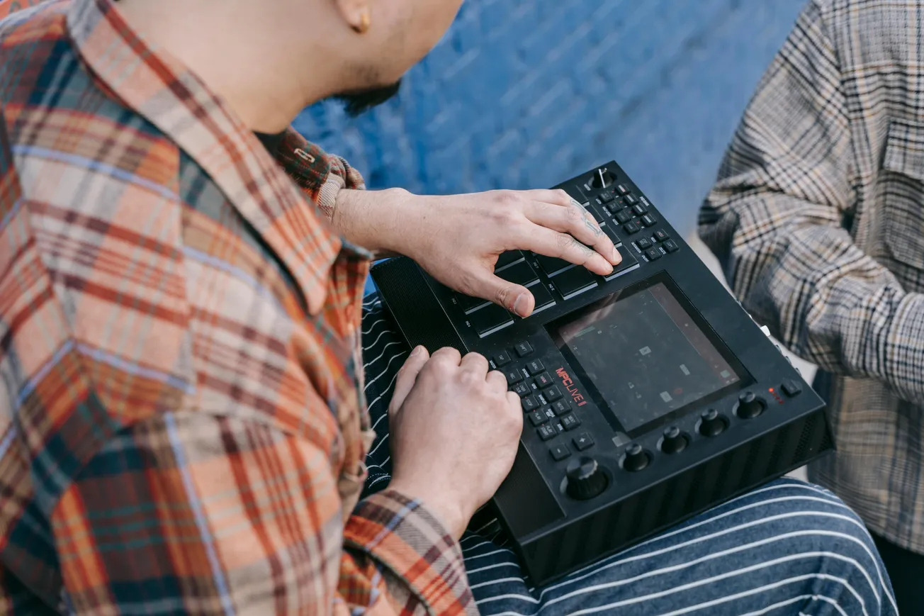 Person in plaid shirt operates a music production device outdoors, fingers pressing buttons on a digital pad, conveying a focused and creative mood.