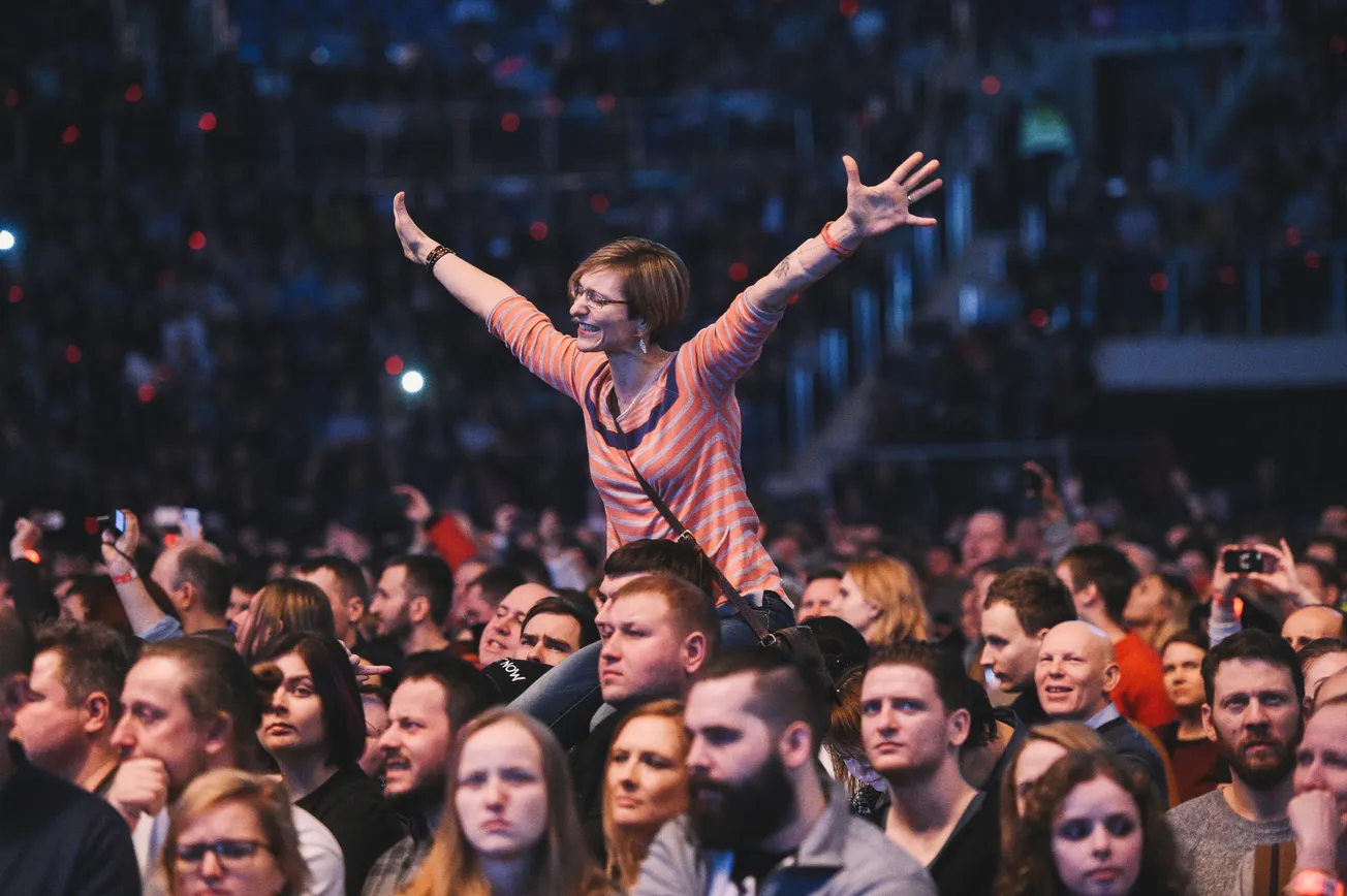 A woman enthusiastically raises her arms, perched on shoulders amidst a dense, lively crowd at a concert. The atmosphere is energetic and celebratory.