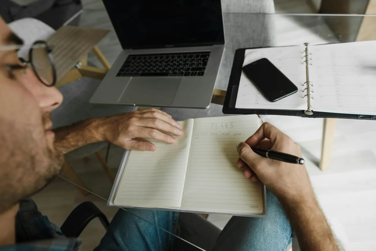 A person writes notes in an open planner on a glass table, surrounded by a laptop and a smartphone. The scene conveys focus and organization.
