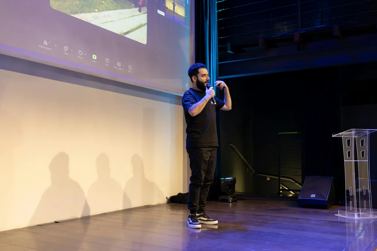 A man in casual attire speaks into a microphone on a stage with a large screen behind him. The setting is modern with a focused and engaging atmosphere.