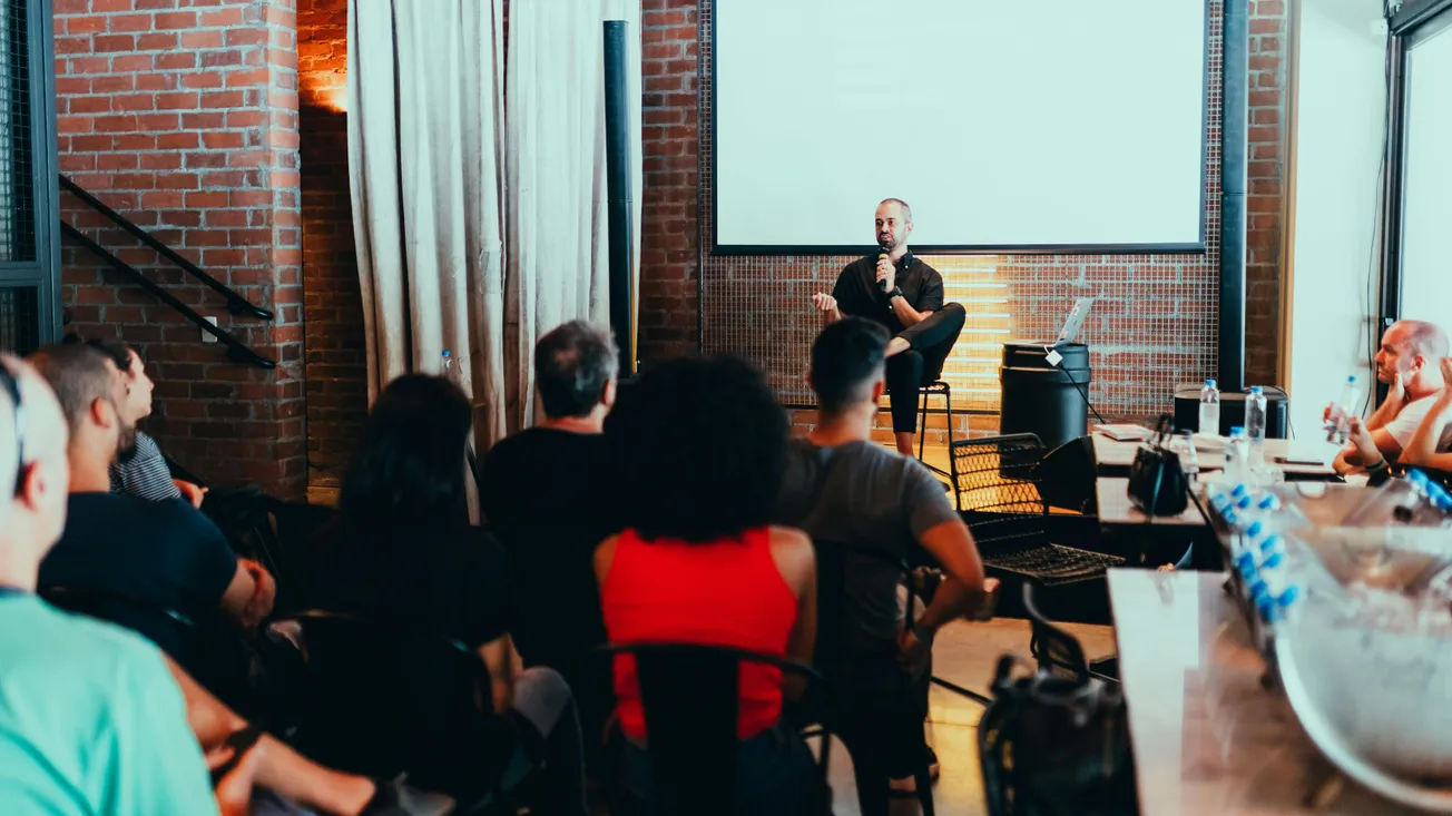 A speaker sits on a stool in front of an audience in a cozy, brick-walled room. The atmosphere is attentive, with viewers focused on the presentation.