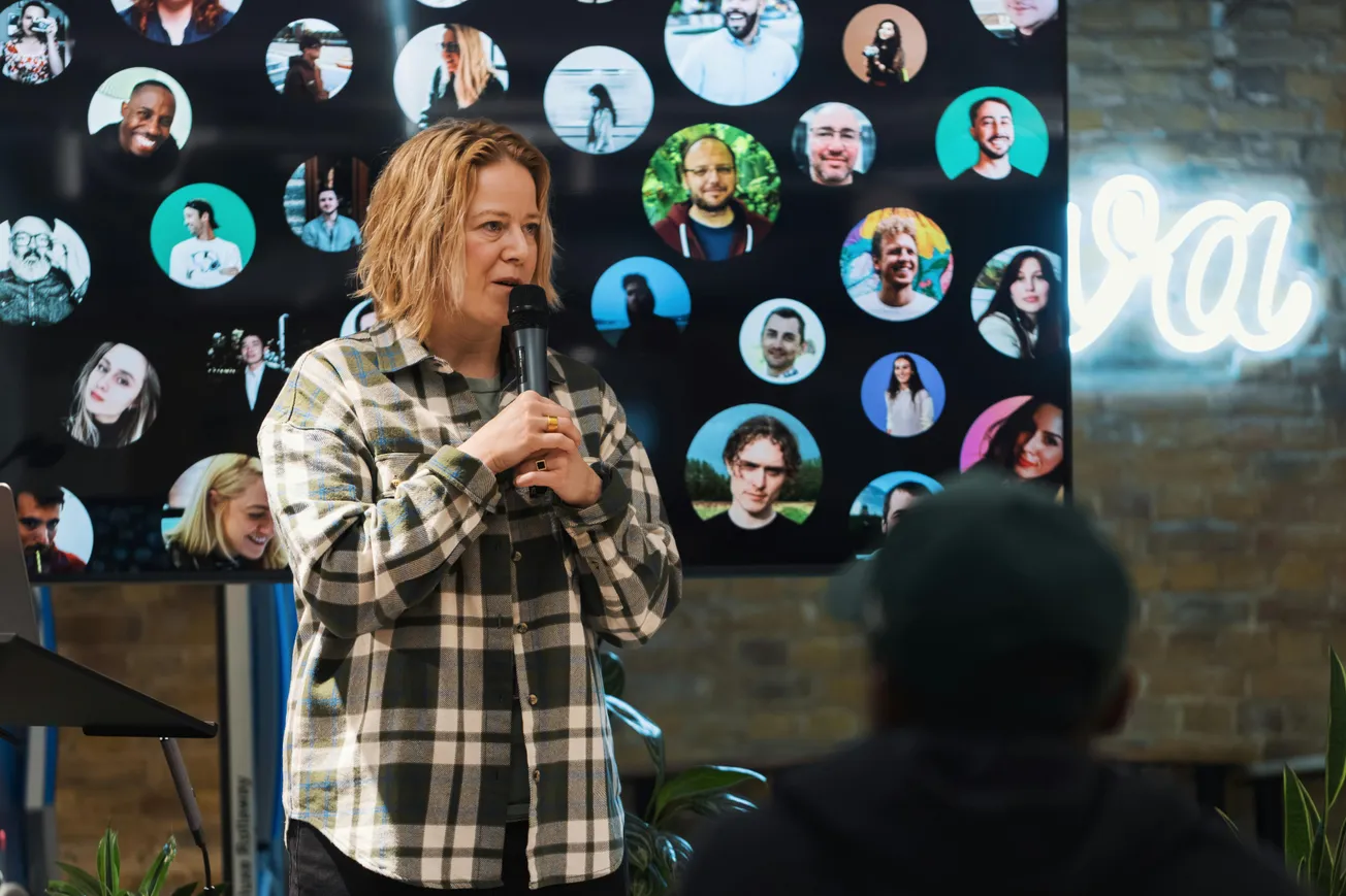 A person speaks into a microphone at an event, wearing a plaid shirt. A screen behind them shows various people's portraits in circles. The setting is casual and lively.