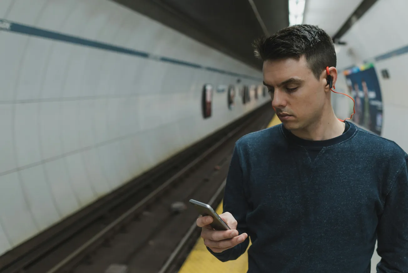 A man with earbuds stands on a subway platform, engrossed in his smartphone. The tunnel is empty, evoking a calm, introspective atmosphere.