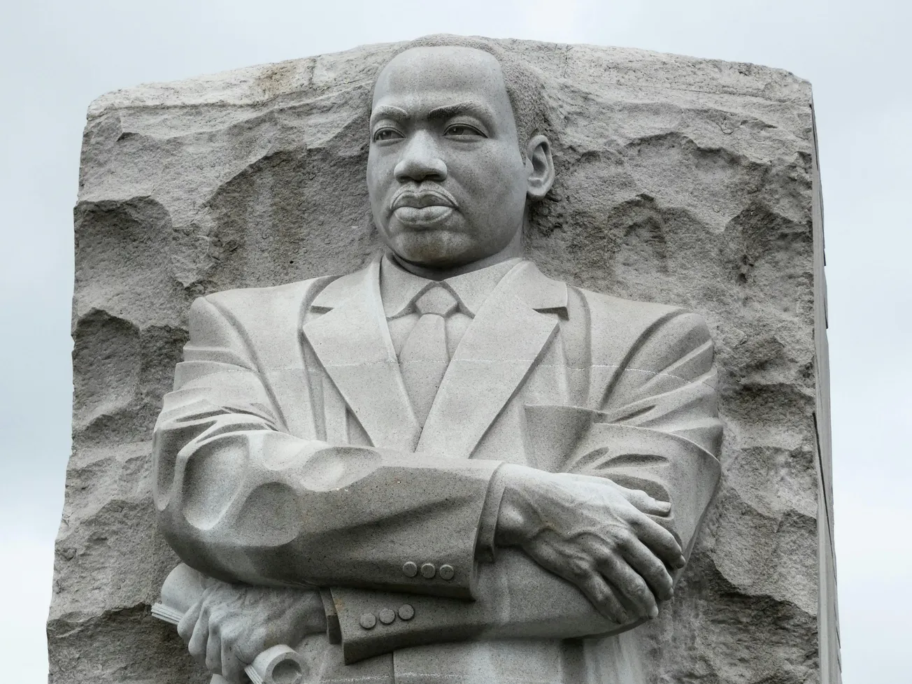 A stone sculpture of MLK in a suit with arms crossed, emerging from a rocky background. His expression is solemn and thoughtful, set against a cloudy sky.