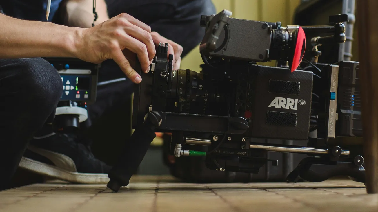 Hands adjust a professional ARRI camera on a movie set. The floor is tiled, and the scene conveys a focused, technical atmosphere.
