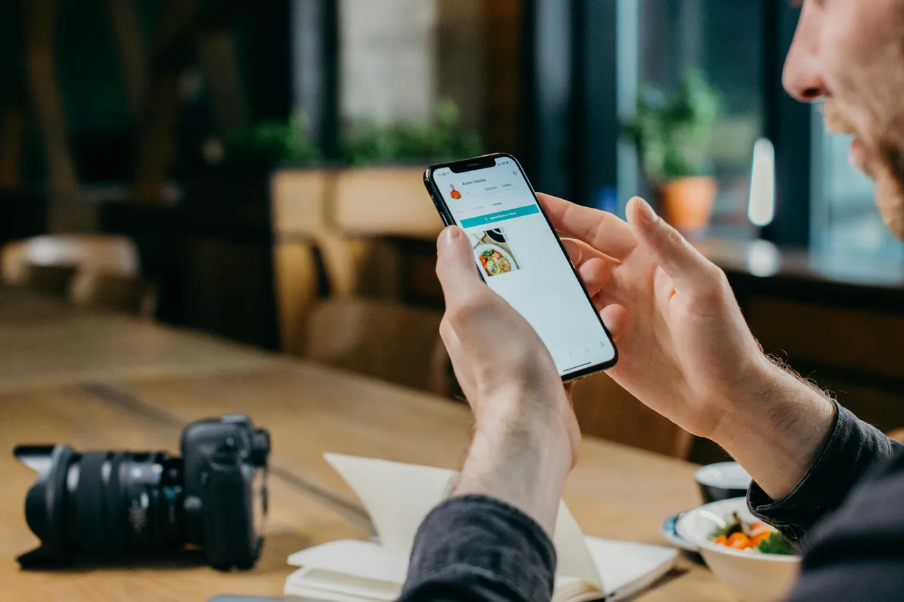 Man in a cafe checks his smartphone, displaying a food delivery app. A camera and notebook lie on the table, suggesting a casual, creative atmosphere.