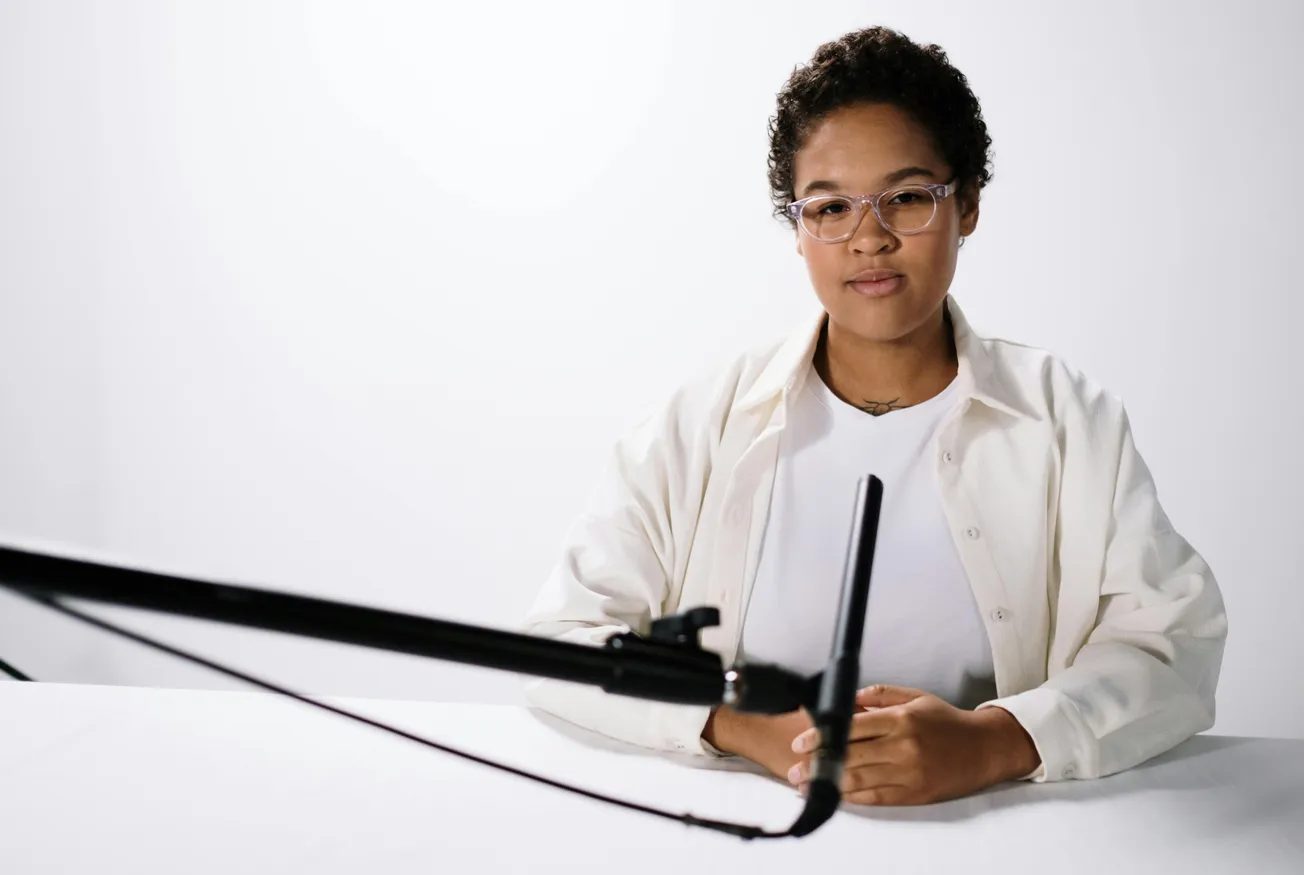 A person with glasses sits at a table against a white background, speaking into a black microphone. The tone is professional and focused.