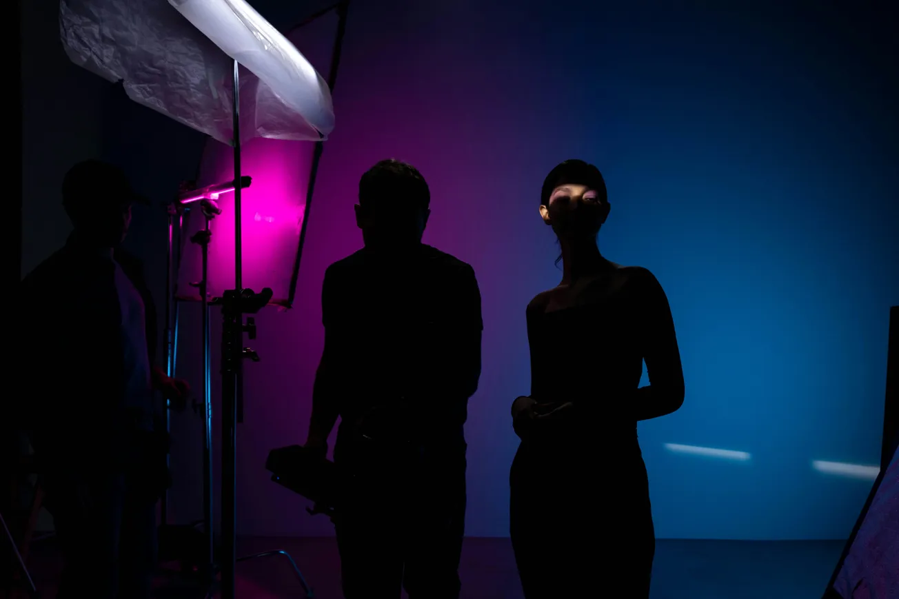 Silhouettes of two individuals in a dimly lit room, with vibrant pink and blue lighting creating a mysterious and artistic atmosphere.
