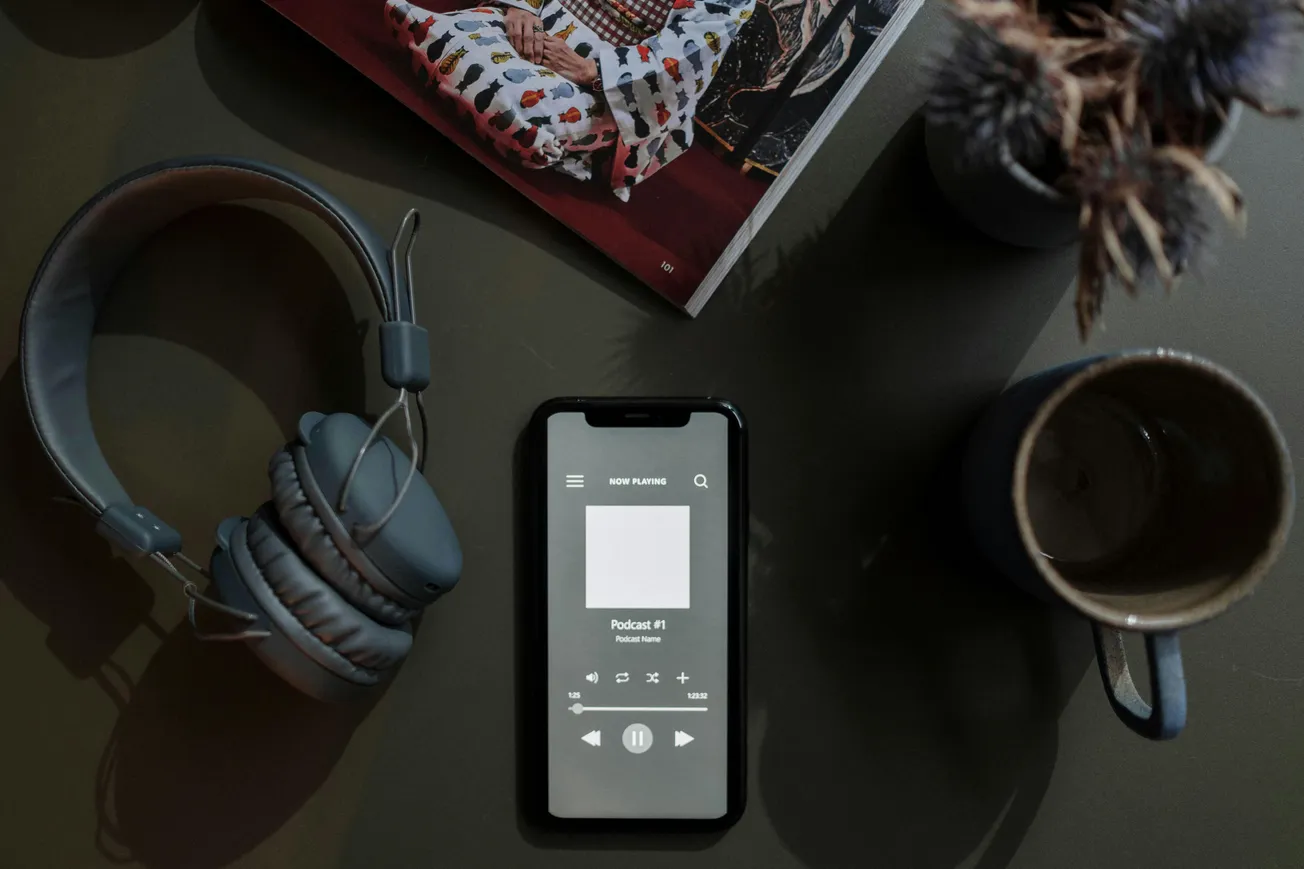 A smartphone displaying a podcast app is placed on a table with gray headphones, a coffee cup, a magazine, and a small plant, creating a cozy and calm atmosphere.