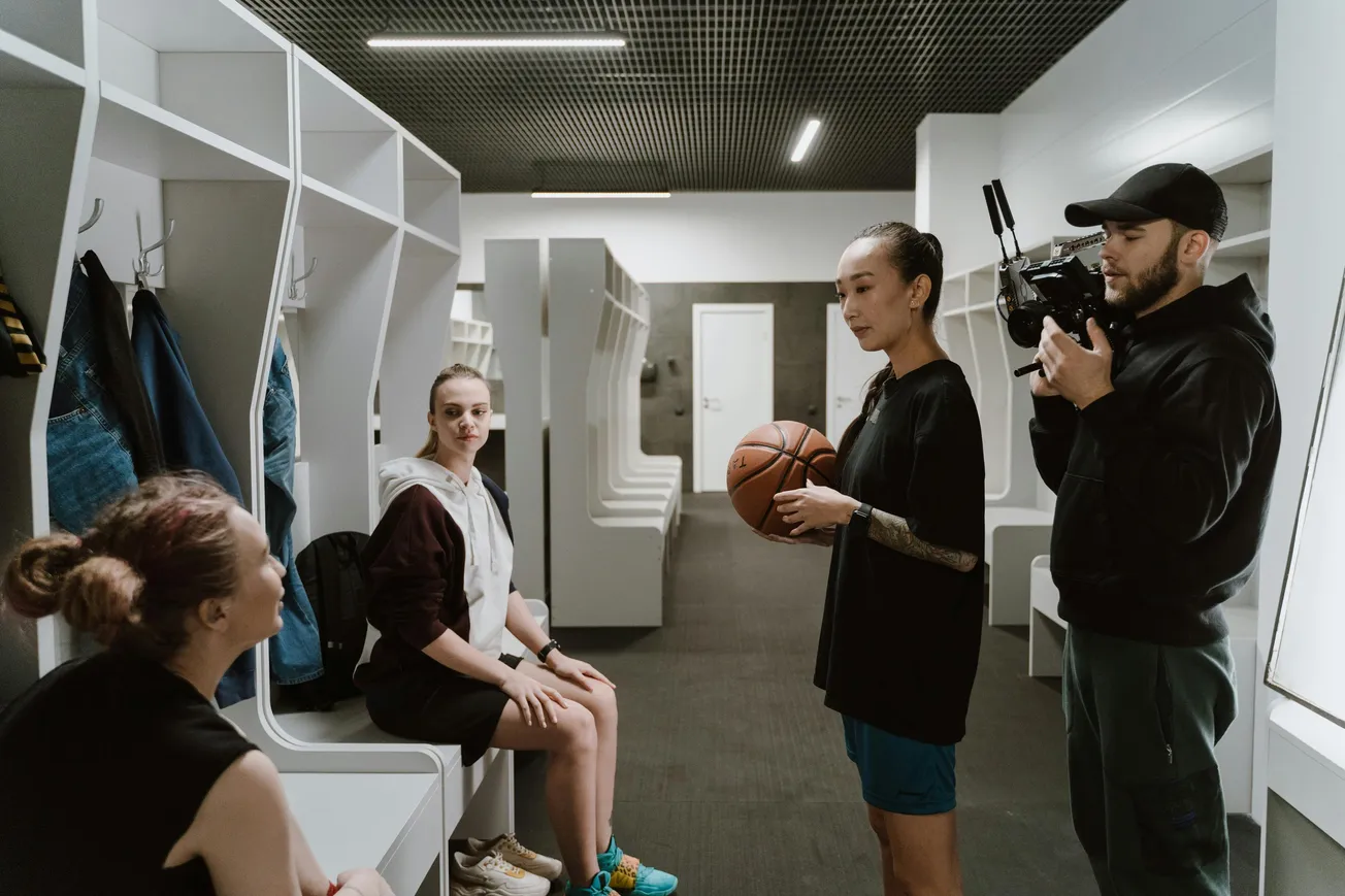 A group of athletes sits in a locker room, engaged in conversation. One holds a basketball, while another person nearby records them with a camera.