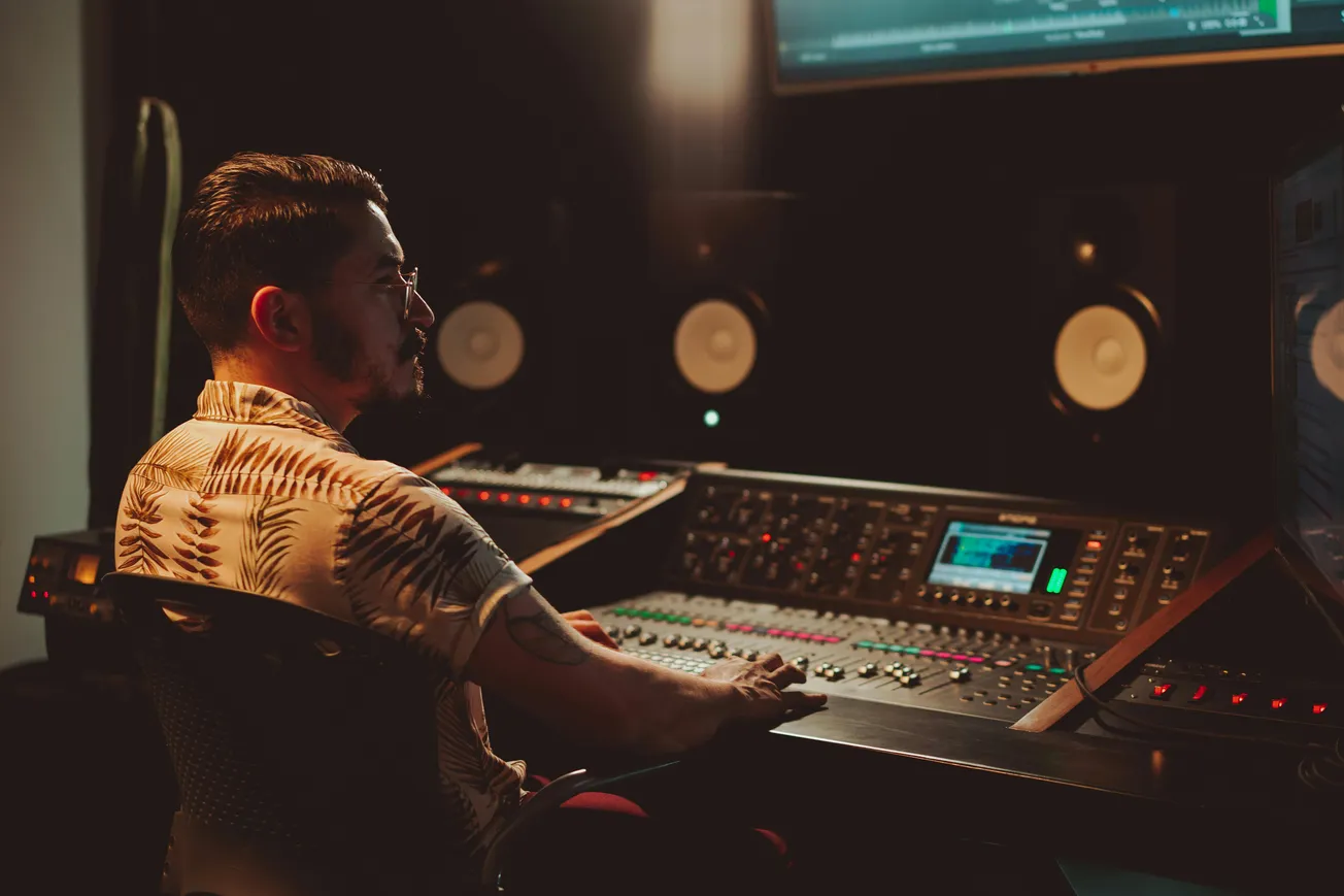 Man in a patterned shirt sits focused at a large audio mixing console in a dimly lit recording studio with speakers and a screen. Calm, creative atmosphere.