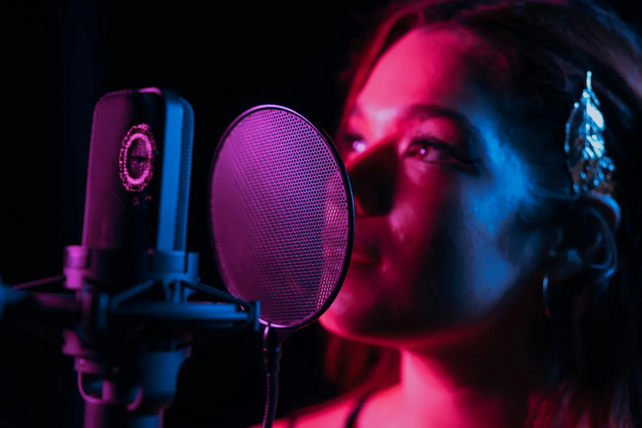 A woman sings into a studio microphone with a pop filter, lit by vibrant pink and blue lights. Her expression is focused and passionate.