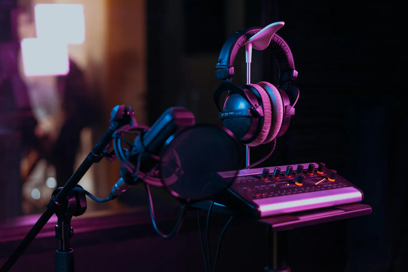 A dimly lit recording studio scene with a microphone and pop filter in the foreground, headphones hanging on a stand, and a mixing console under soft purple light.