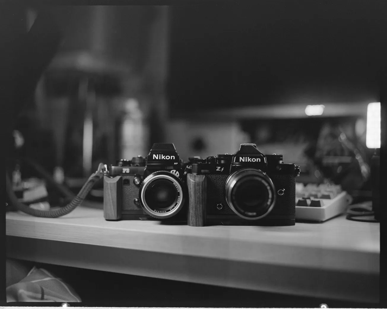 Black and white photo of two Nikon cameras on a wooden desk, surrounded by cables and equipment. The scene conveys a nostalgic, vintage feel.