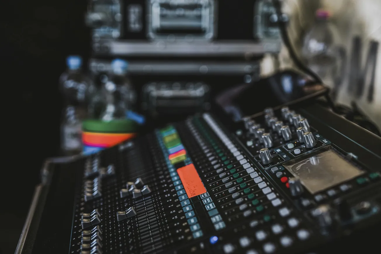 Close-up of a sound mixing board with sliders, knobs, and colorful labels. In the background, blurred water bottles and a flight case are visible.
