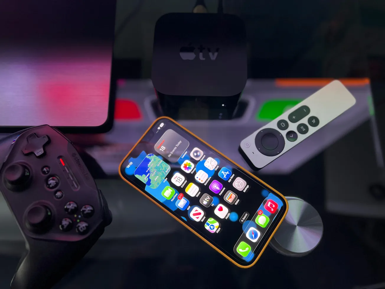 A smartphone with app icons is on a reflective surface beside a game controller, a TV remote, and an Apple TV box, conveying a tech-focused atmosphere.