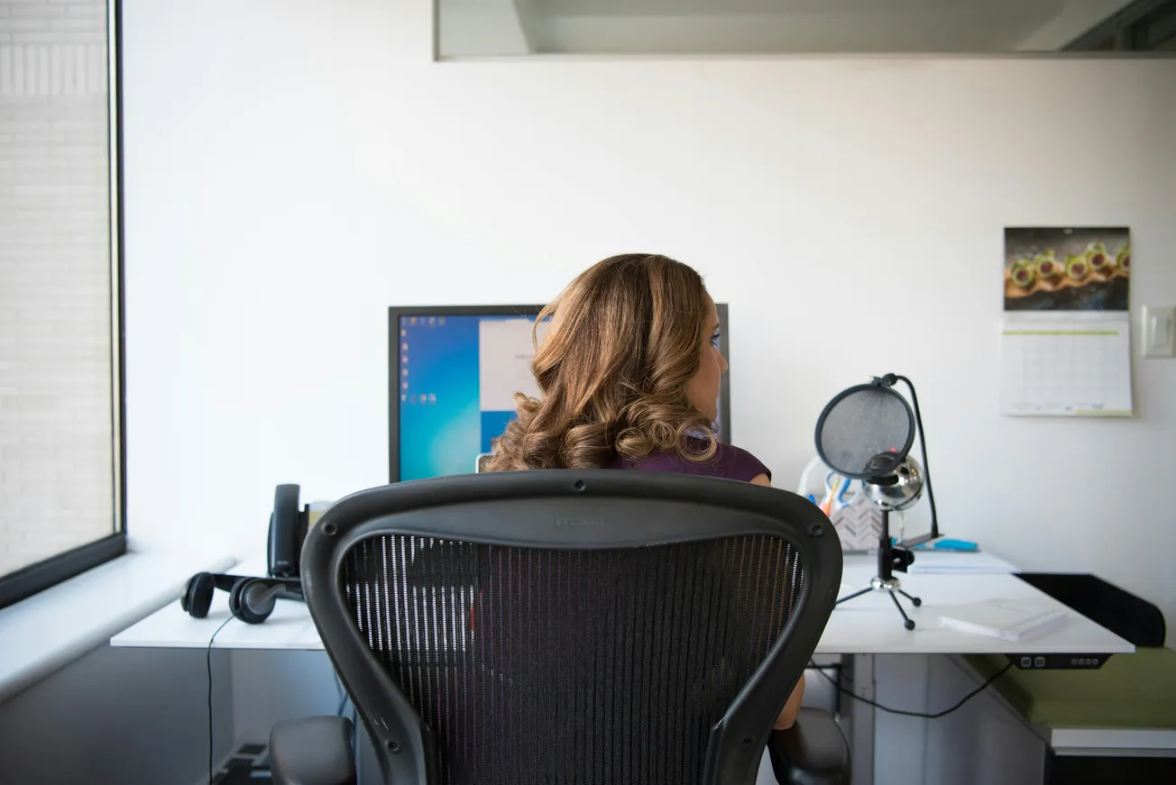 A woman with wavy hair sits in an office chair, facing a computer on a white desk. Nearby are a microphone, calendar, and window, conveying a workspace setting.