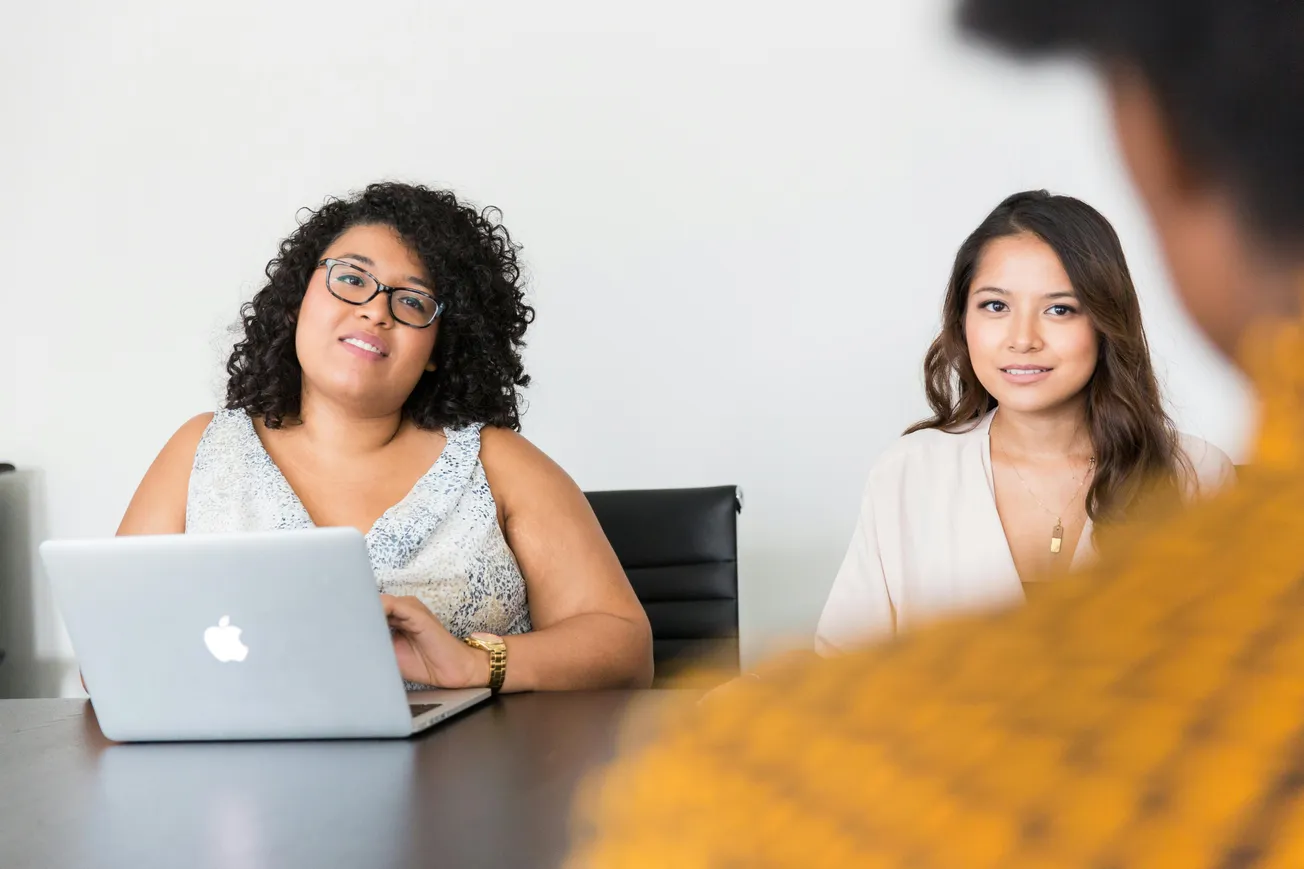 Two women sitting at a table in a meeting, one with a laptop. They appear engaged, listening attentively. The atmosphere is professional and focused.