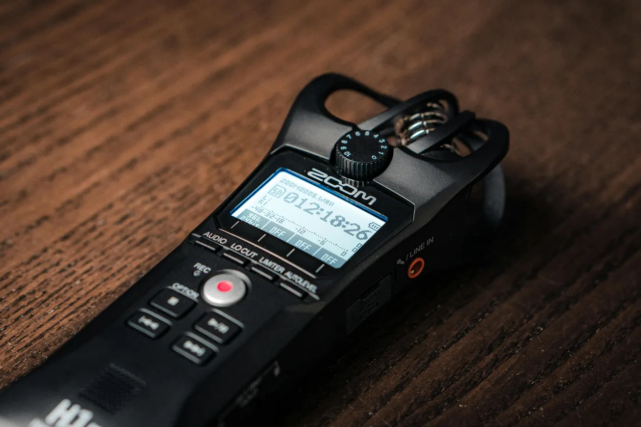 Close-up of a portable audio recorder on a wooden surface. The device displays recording time and has various buttons and a prominent red recording button.