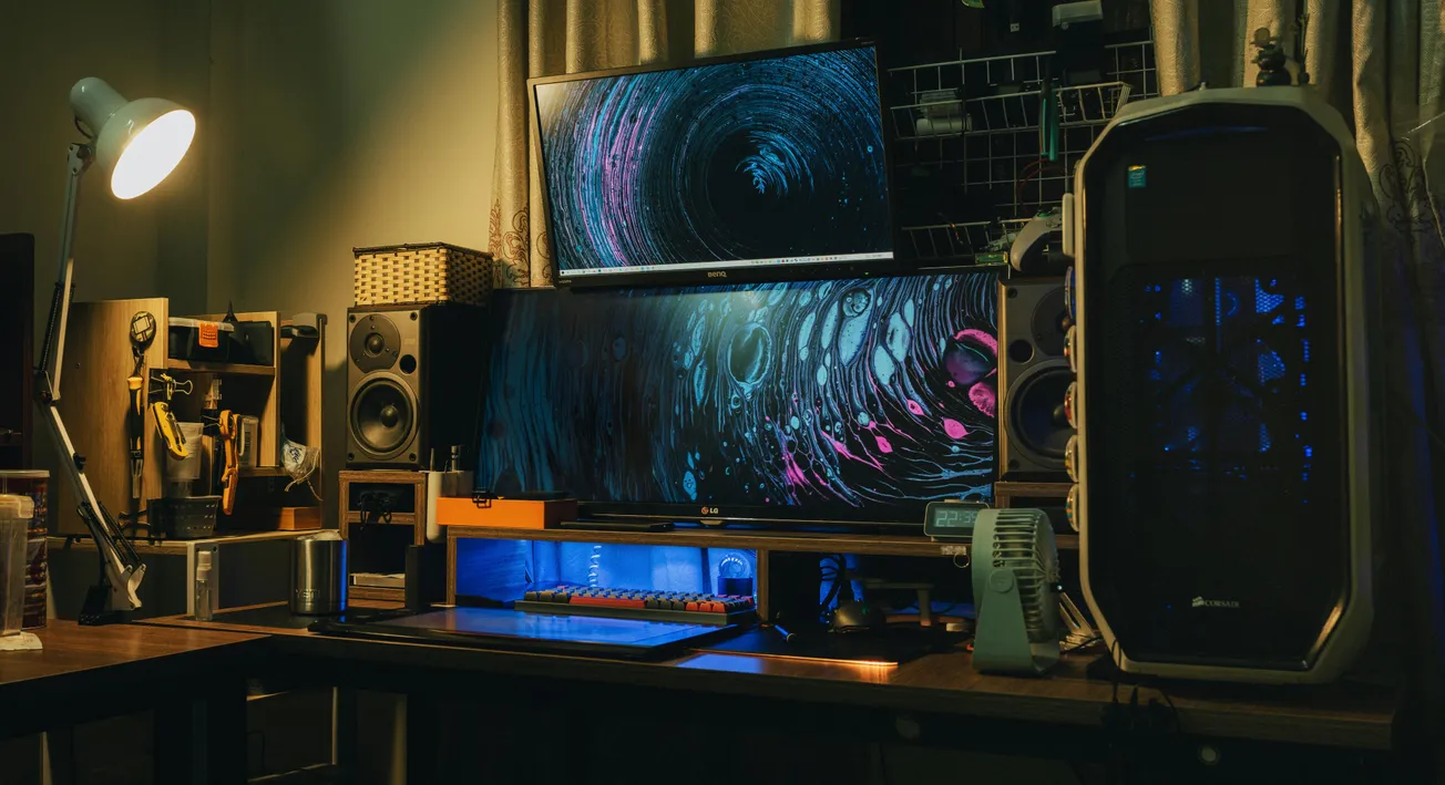 Cozy workspace with dark, ambient lighting features dual monitors displaying abstract art, a gaming PC with blue lighting, speakers, and a desk lamp.