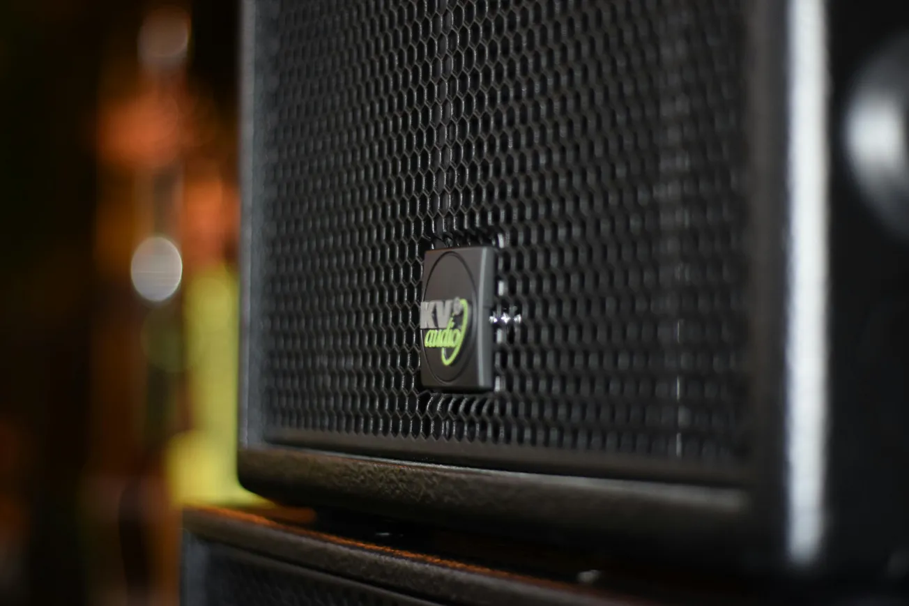 Close-up of a textured black speaker grill with a green logo at the center, set against a blurred, warm-toned background. The scene feels musical and dynamic.