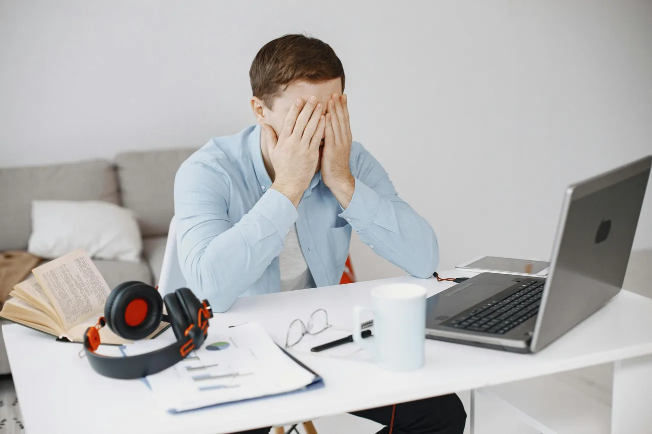 A man in a blue shirt sits at a desk, covering his face in frustration. A laptop, headphones, open book, and documents are scattered on the table, conveying stress.
